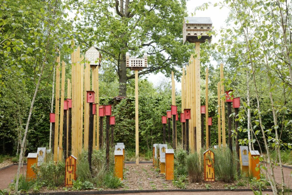 Photo d'un jardin avec tiges de bois plantées dans le sol, sur lesquelles sont accrochées des cabanes à oiseaux de tailles variées. Oeuvre intitulée Pollinators city, Festival International des Jardins, 2024 © E. Sander