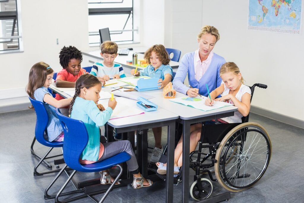 Handicap à l'école - une institutrice aide des enfants autour d'une table, une est en fauteuil roulant.